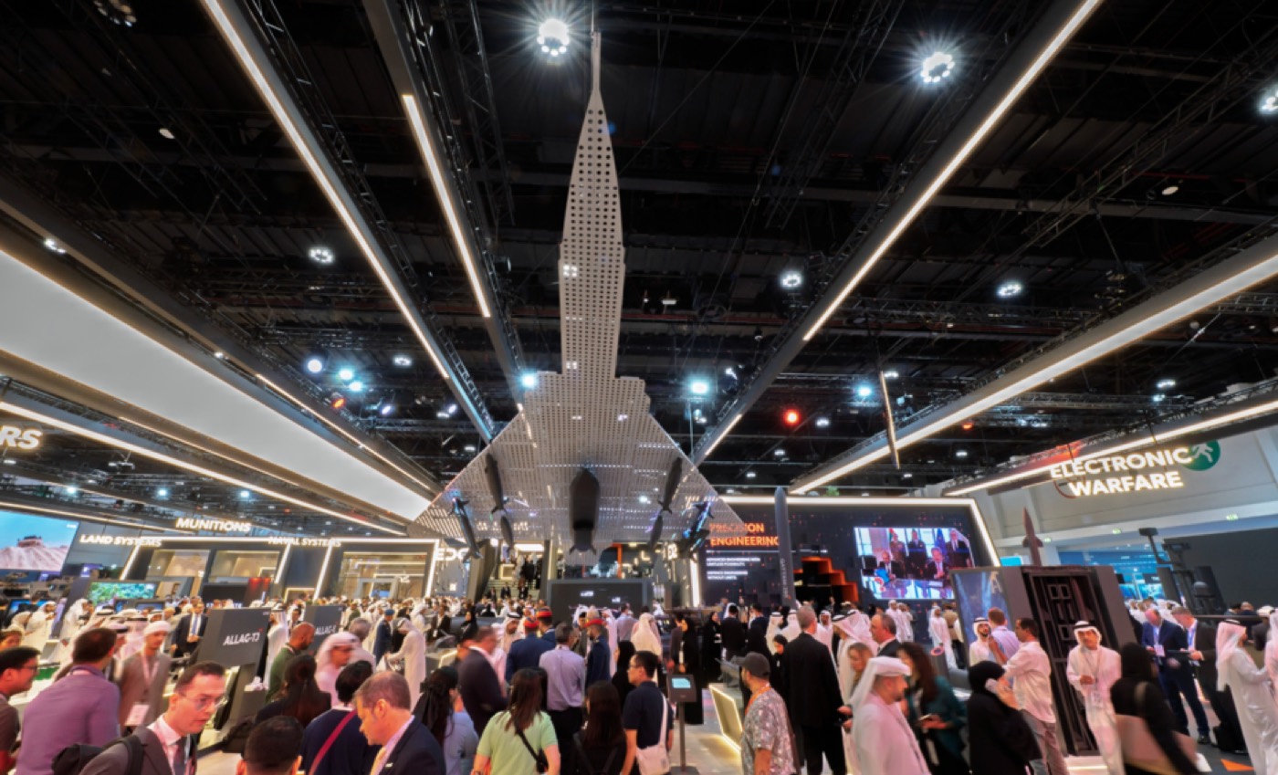 The main exhibition hall at IDEX, أدنيك أبوظبي, with a full-size missile suspended above a crowded floor and the Electronic Warfare and Land Systems halls behind
